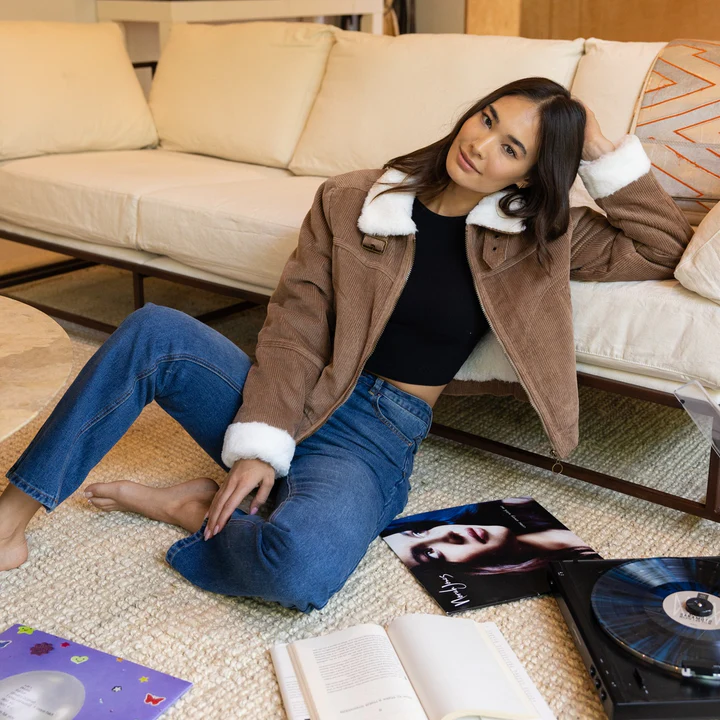 A woman at home wearing dark blue jeans, a black tee, and a corduroy jacket